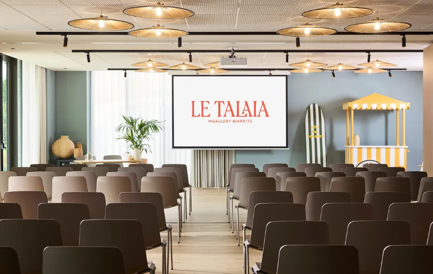Grande salle de conférence de l'hôtel Le Talaia MGallery Biarritz en configuration théâtre, avec rangées de chaises taupe, plafond orné de suspensions en rotin, écran de projection, planche de surf rayée et stand de rafraîchissements rayé jaune et blanc en décoration.