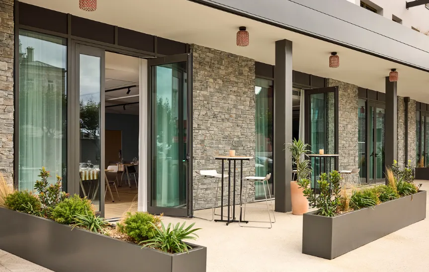 Terrasse de pause séminaire de l'hôtel Le Talaia, avec façade en pierre naturelle, baies vitrées pliantes ouvertes sur la salle intérieure dressée pour un coffee break, tables hautes et jardinières anthracite végétalisées.