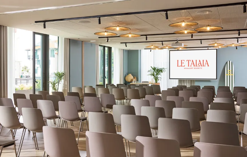 Salle de conférence de l'hôtel Le Talaia MGallery Biarritz vue en angle, en configuration théâtre avec nombreuses chaises taupe, grandes fenêtres lumineuses donnant sur l'extérieur, plafond à suspensions en rotin et écran de projection avec logo.