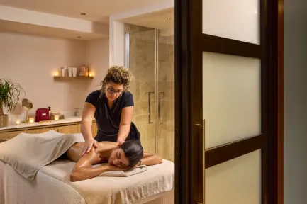 Thérapeute aux cheveux bouclés massant le dos d'une cliente allongée sur une table de soin, dans une cabine de spa aux tons beiges chauds éclairée par des bougies, vue depuis l'encadrement d'une porte.