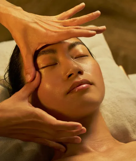 Femme allongée les yeux fermés recevant un soin du visage, avec des mains effectuant un massage au niveau du front et de la mâchoire, dans une ambiance lumineuse aux tons chauds dorés.