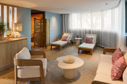 Salle de détente d'un spa avec fauteuils en bois clair, chaises longues, table basse ronde en pierre, murs bleu gris, paravent en bois sculpté et lumière naturelle tamisée par des rideaux blancs.