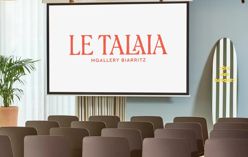 Salle de séminaire lumineuse de l'hôtel Le Talaia MGallery Biarritz en configuration théâtre, avec rangées de chaises taupe face à un écran de projection, planche de surf rayée verte estampillée « Côte des Basques » en décoration et plante tropicale.