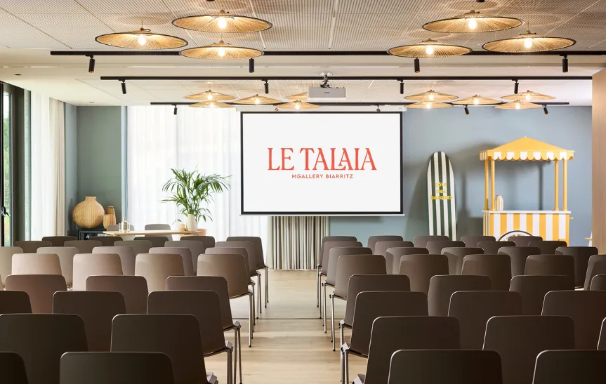 Salle de conférence Talaia de l'hôtel MGallery Biarritz en configuration plénière, vue depuis le fond de la salle, avec rangées de chaises taupe, plafond à suspensions en rotin, écran de projection affichant le logo, plante tropicale et stand de limonade rayé jaune.