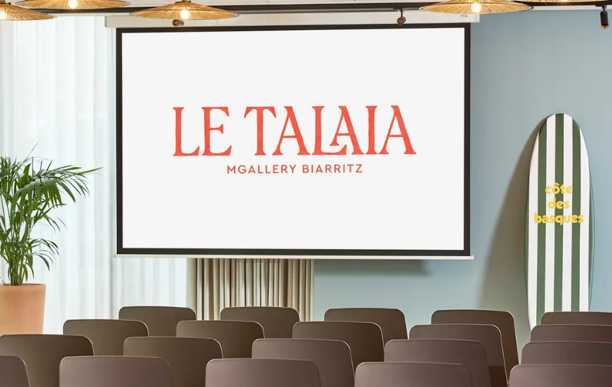 Salle de conférence de l'hôtel Le Talaia MGallery Biarritz avec rangées de chaises taupe, écran de projection affichant le logo de l'établissement en rouge, plante tropicale et planche de surf rayée verte et blanche en décoration.