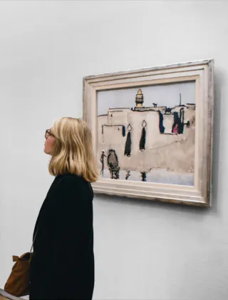 Une femme blonde à lunettes contemplant un tableau encadré représentant une scène de rue orientale avec personnages, exposé sur un mur blanc de galerie ou de musée.
