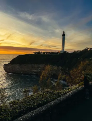 Le phare de Biarritz au coucher du soleil, dressé sur une falaise rocheuse dominant l'océan Atlantique, sous un ciel aux dégradés de bleu, orange et doré