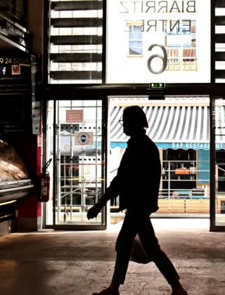 Silhouette d'une personne marchant dans les Halles de Biarritz, avec en arrière-plan la devanture lumineuse du marché couvert et l'enseigne « Biarritz Entre » visible en reflet sur la vitrine.