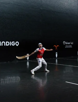 Joueur de pelote basque en tenue rouge et blanche effectuant un geste de frappe à la chistera dans un fronton couvert aux murs noirs, avec les logos des sponsors Indigo et Bien'ici visibles en arrière-plan.
