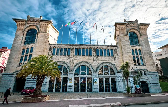 Façade du Casino Municipal de Biarritz, bâtiment Belle Époque en pierre de taille avec grandes arcades vitrées, deux tours encadrantes et drapeaux internationaux, entouré de palmiers sous un ciel nuageux.