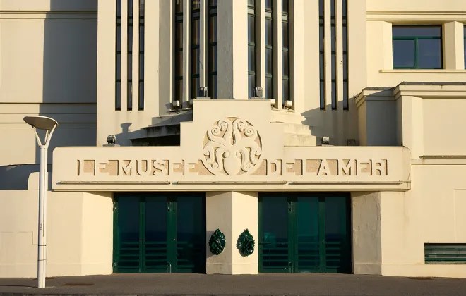 Façade Art déco du Musée de la Mer de Biarritz, avec son inscription en lettres dorées en relief, ses grandes portes vitrées vert émeraude et ses ornements sculptés, baignée d'une lumière dorée rasante.