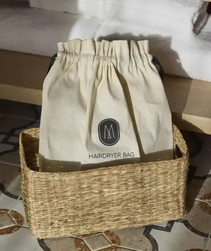 Pochette en coton écru marquée "Hairdryer Bag" posée dans un panier tressé, sur un carrelage à motifs, avec des serviettes blanches en arrière-plan.