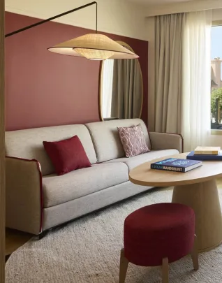 Salon d'une chambre d'hôtel élégante avec un canapé beige, une table basse ronde en bois, un fauteuil et un pouf bordeaux, une grande fenêtre offrant une vue sur la mer, dans une décoration aux tons chauds rose et crème