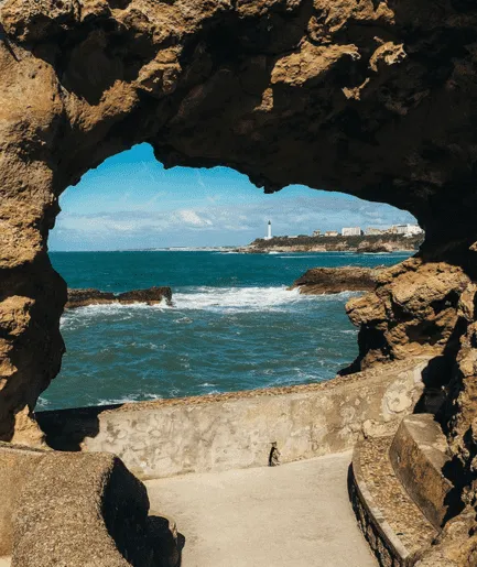 Vue encadrée par une arche rocheuse naturelle sur la côte sauvage de Biarritz, avec l'océan agité et le phare en arrière-plan.