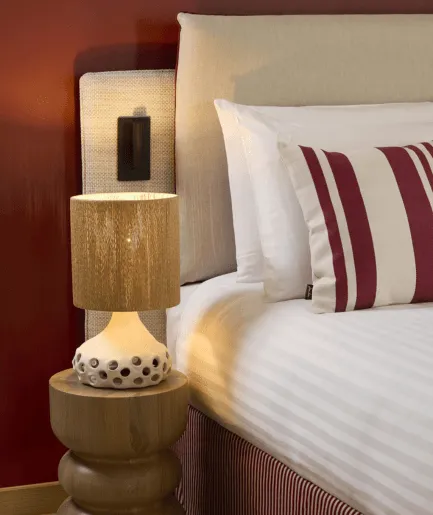 Détail de chevet avec lampe en céramique ajourée sur table de nuit en bois tourné, lit avec coussins rayés bordeaux et tête de lit en lin sur mur rouge.