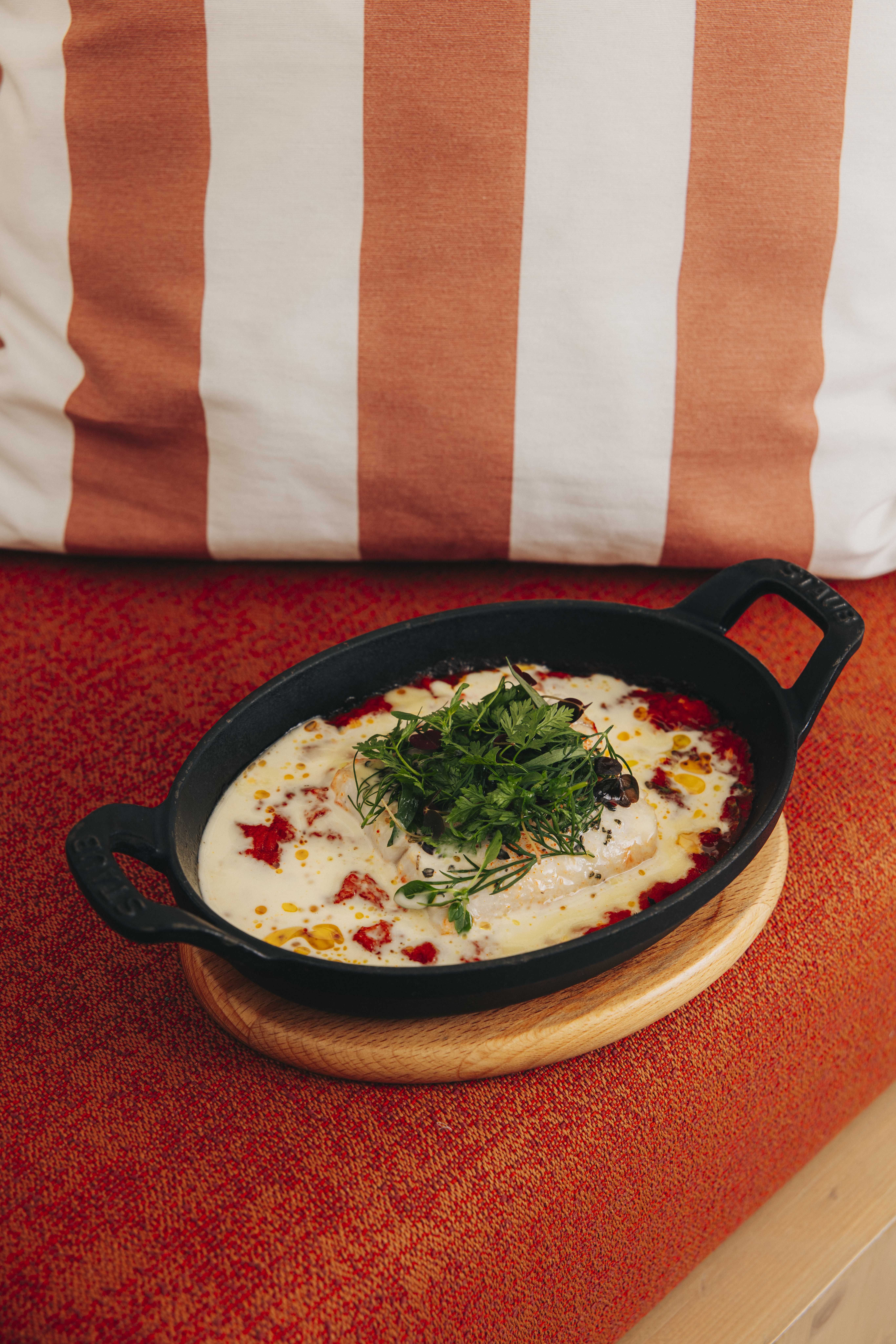 Main tenant une fourchette au-dessus d'un plat cuisiné servi en cocotte noire ovale, garni de fromage fondu, sauce tomate et herbes fraîches, posé sur une table en bois