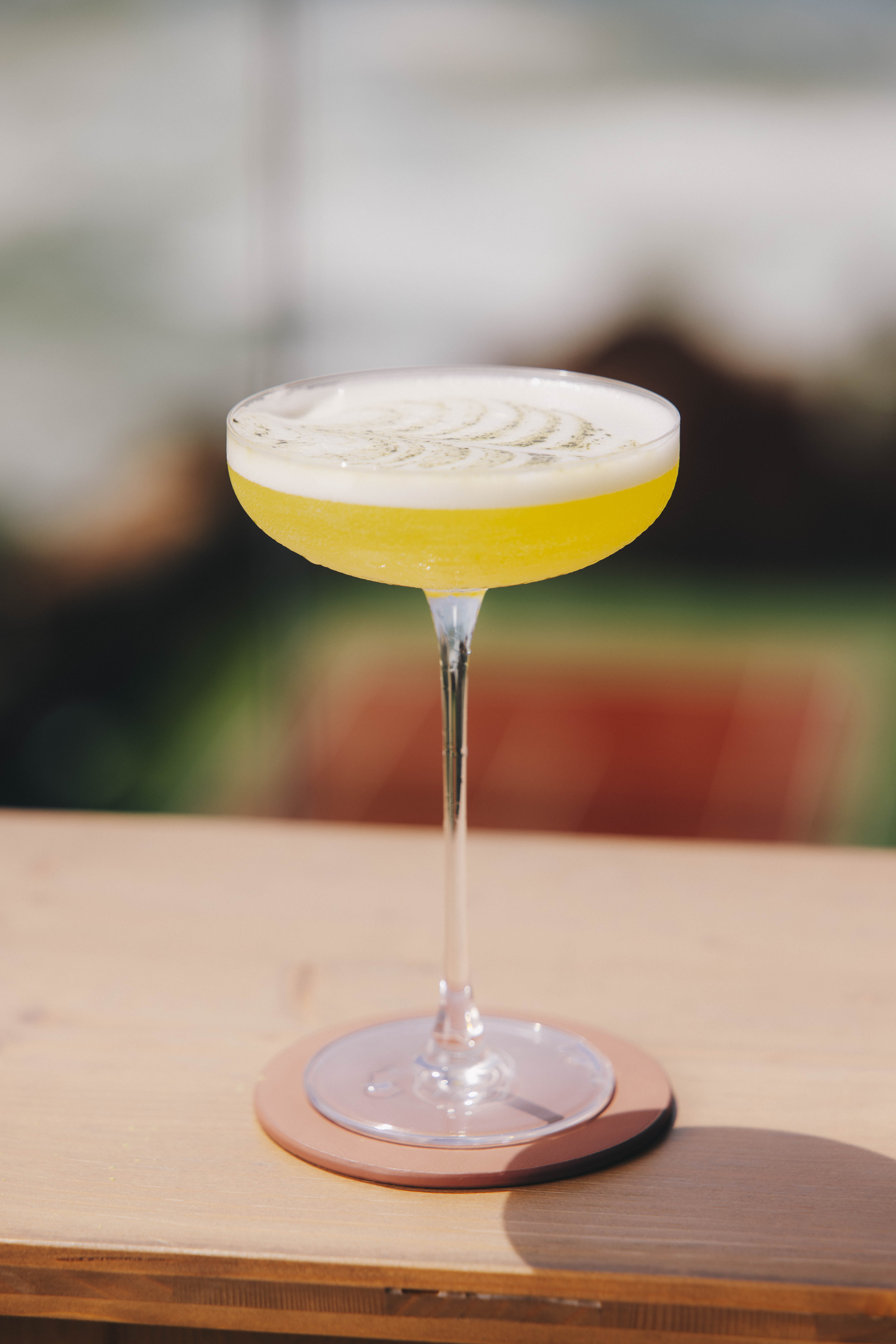 Cocktail glacé servi dans un verre taillé avec une paille noire et saupoudré d'épices dorées, posé sur une carte de menu rose fuchsia sur une table en bois, devant un bouquet de fleurs roses fuchsia en arrière-plan