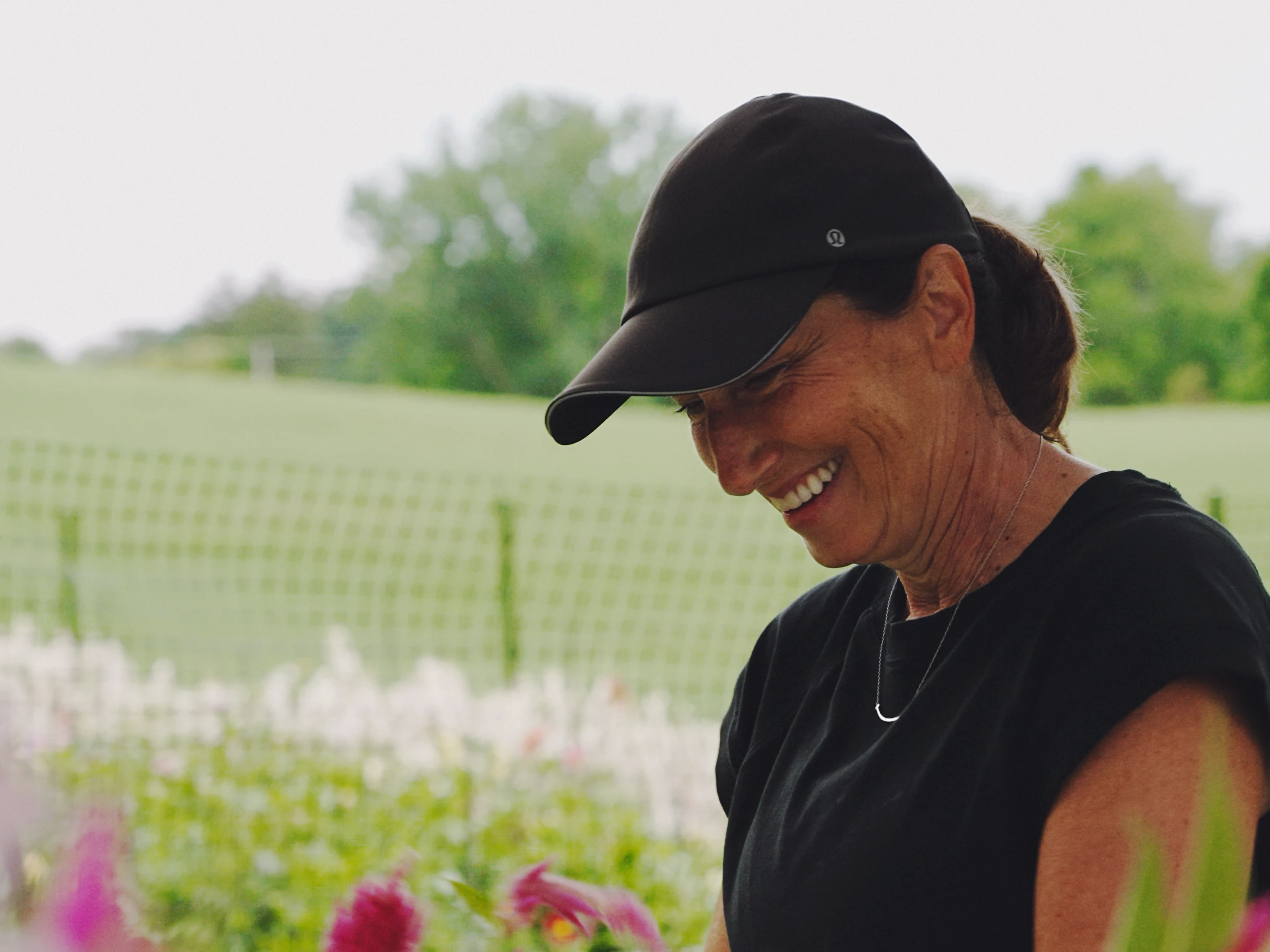 A woman smiling in the flower field, rows of blooms and fencing behind her.