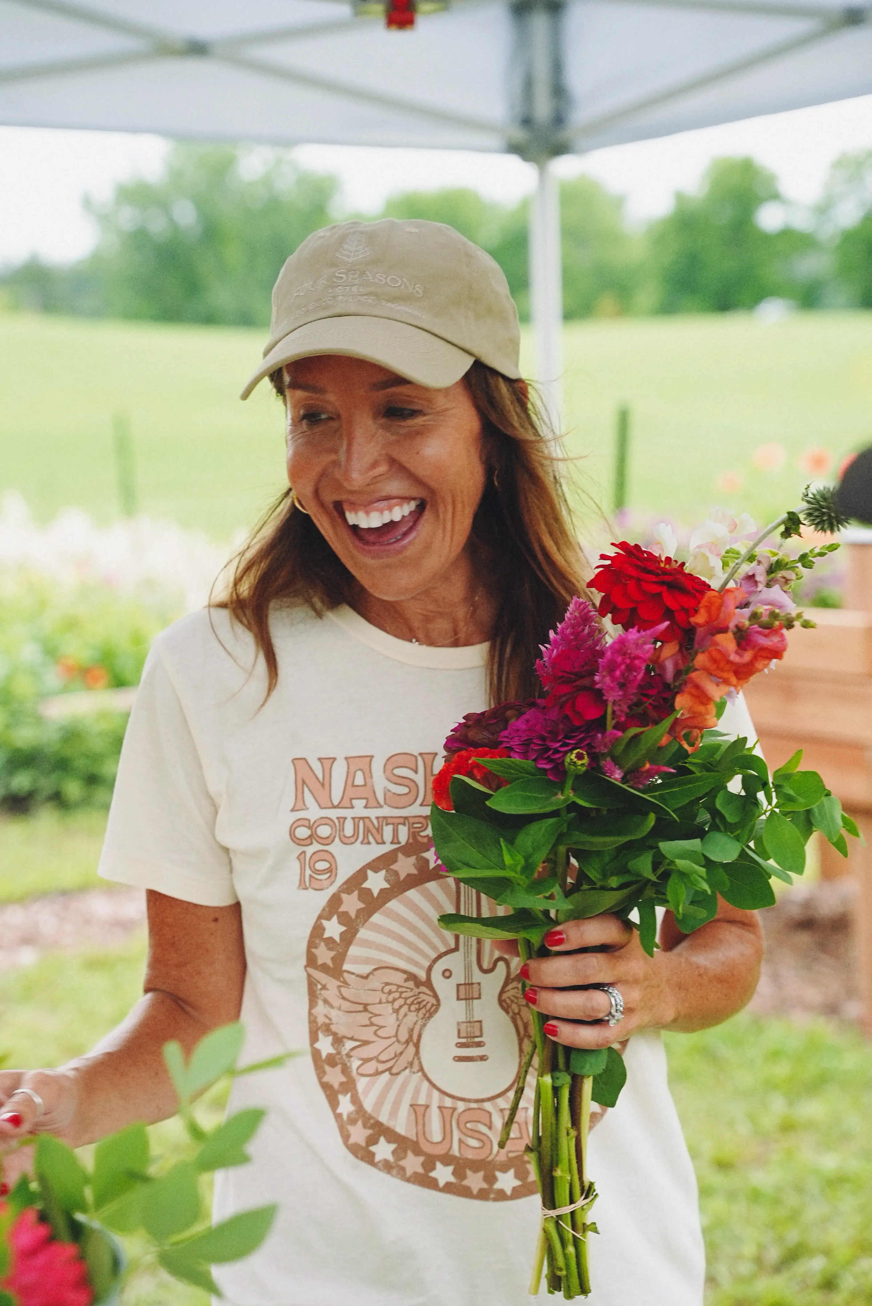 A woman laughing and holding a bouquet of red, pink, and orange flowers at an outdoor market stand.