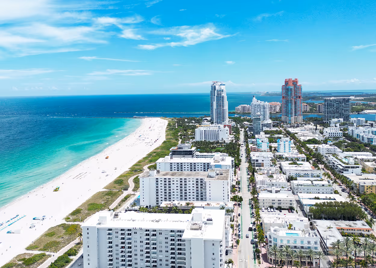 Aerial view of a coastal city with tall buildings, a white sandy beach, and the ocean under a blue sky.
