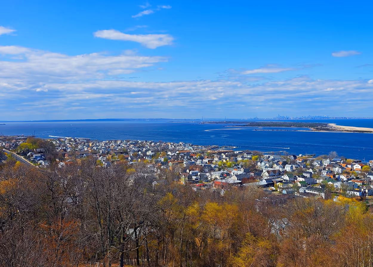 Panoramic view over a coastal town with houses, trees, and a large body of water under a partly cloudy blue sky.