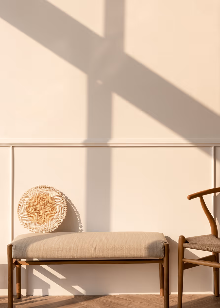 Minimalist interior with beige cushioned bench, woven straw round wall decor, and wooden chair against a light wall with sunlight shadows.