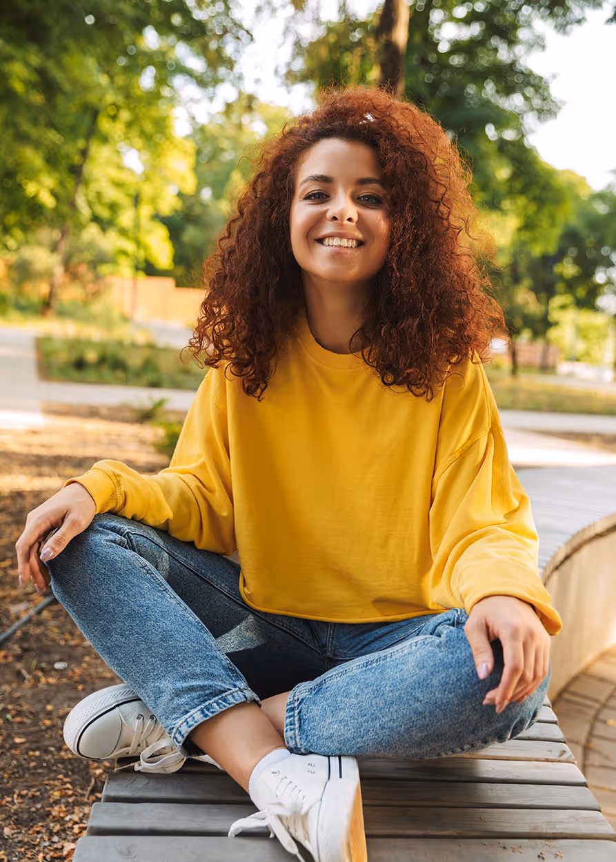 Young woman with curly red hair wearing a yellow sweatshirt and jeans sitting cross-legged on a wooden bench in a park.