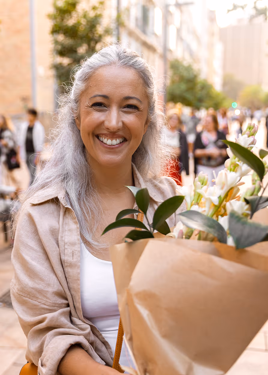 Smiling woman with gray hair holding a bouquet of white flowers wrapped in brown paper on a city street.