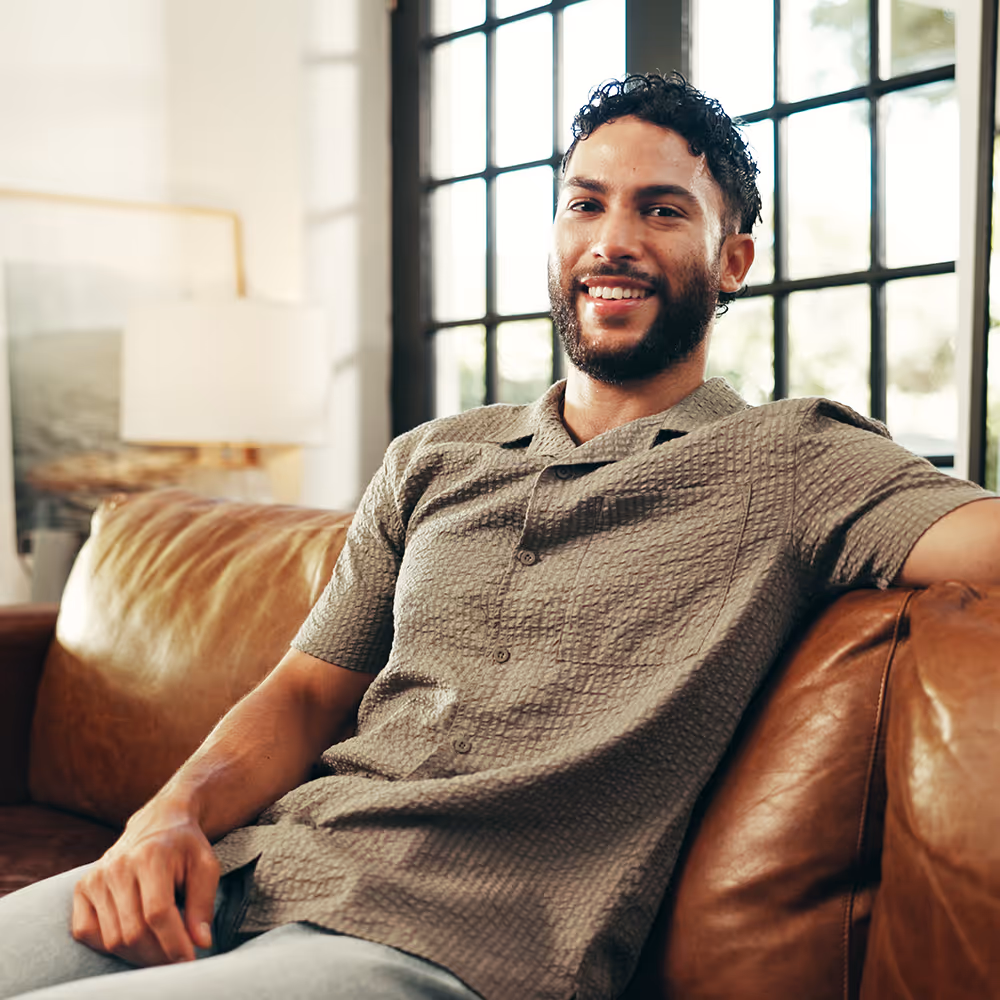 Smiling man with curly hair and beard sitting relaxed on a brown leather couch in a sunlit room with large windows.