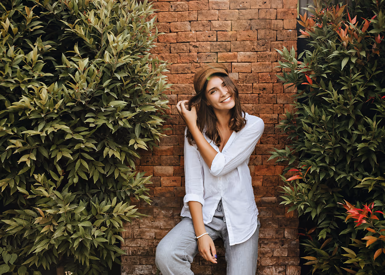 Smiling woman wearing a brown cap and white shirt sitting against a brick wall surrounded by green foliage.