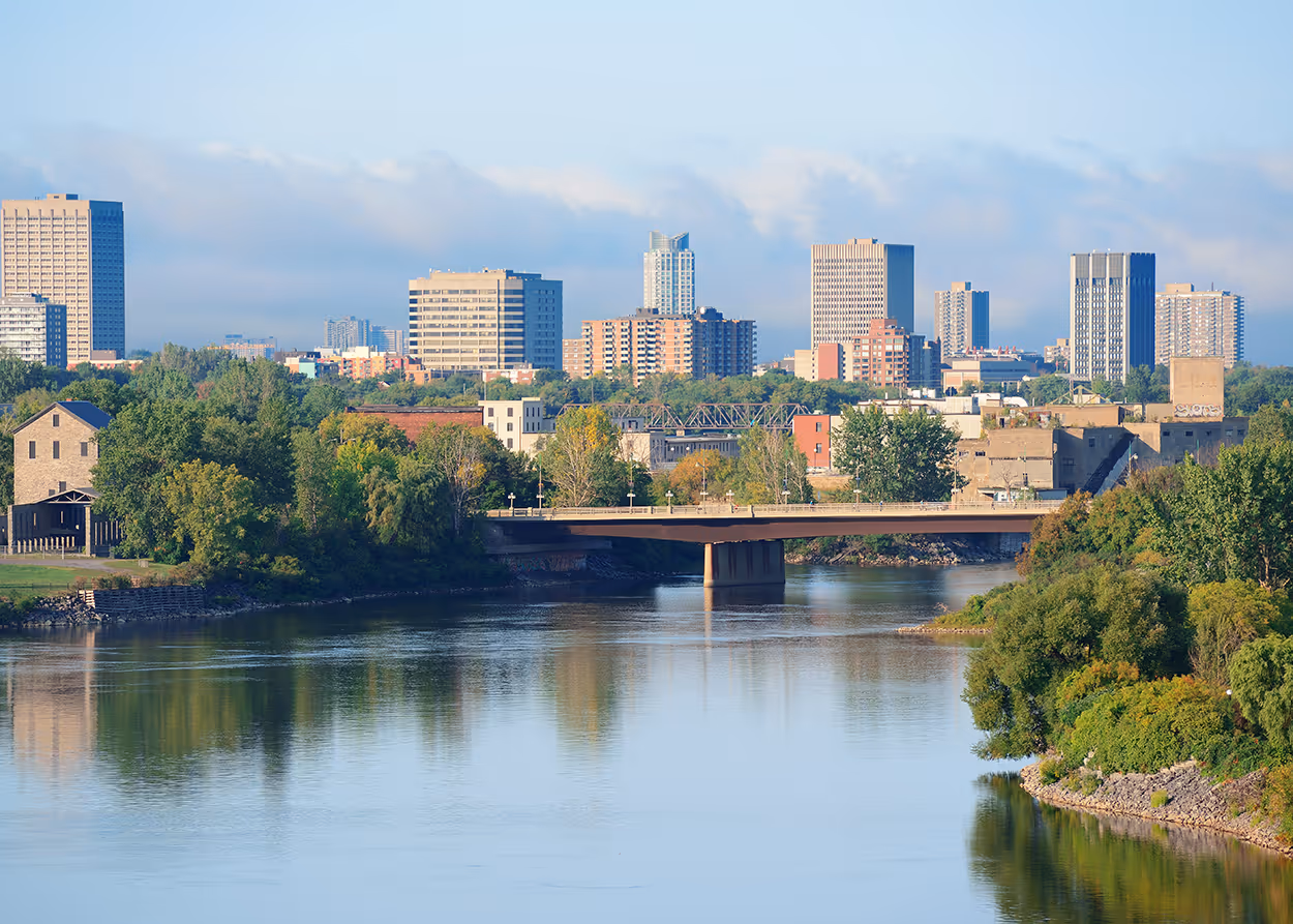 City skyline with modern and older buildings behind a river and a bridge surrounded by green trees on a clear day.