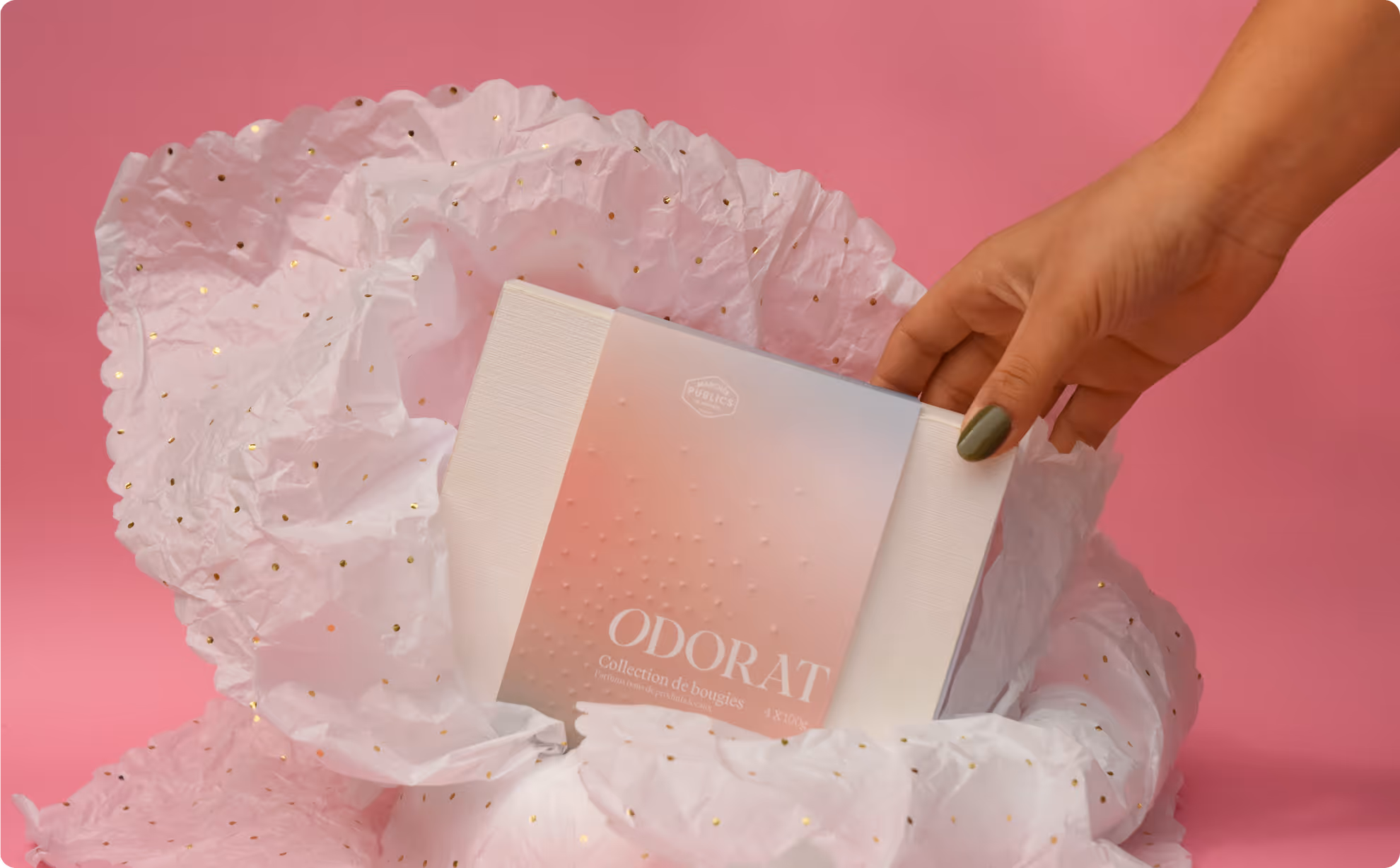 Hand with green nail polish holding a boxed candle collection labeled Odorat on pink background. Credit: May-lee Abel