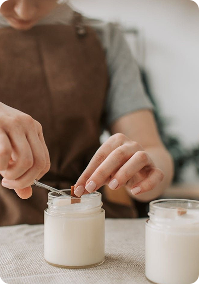 Person placing a wooden wick into a white candle jar while wearing a brown apron. Credit: Chatgpt