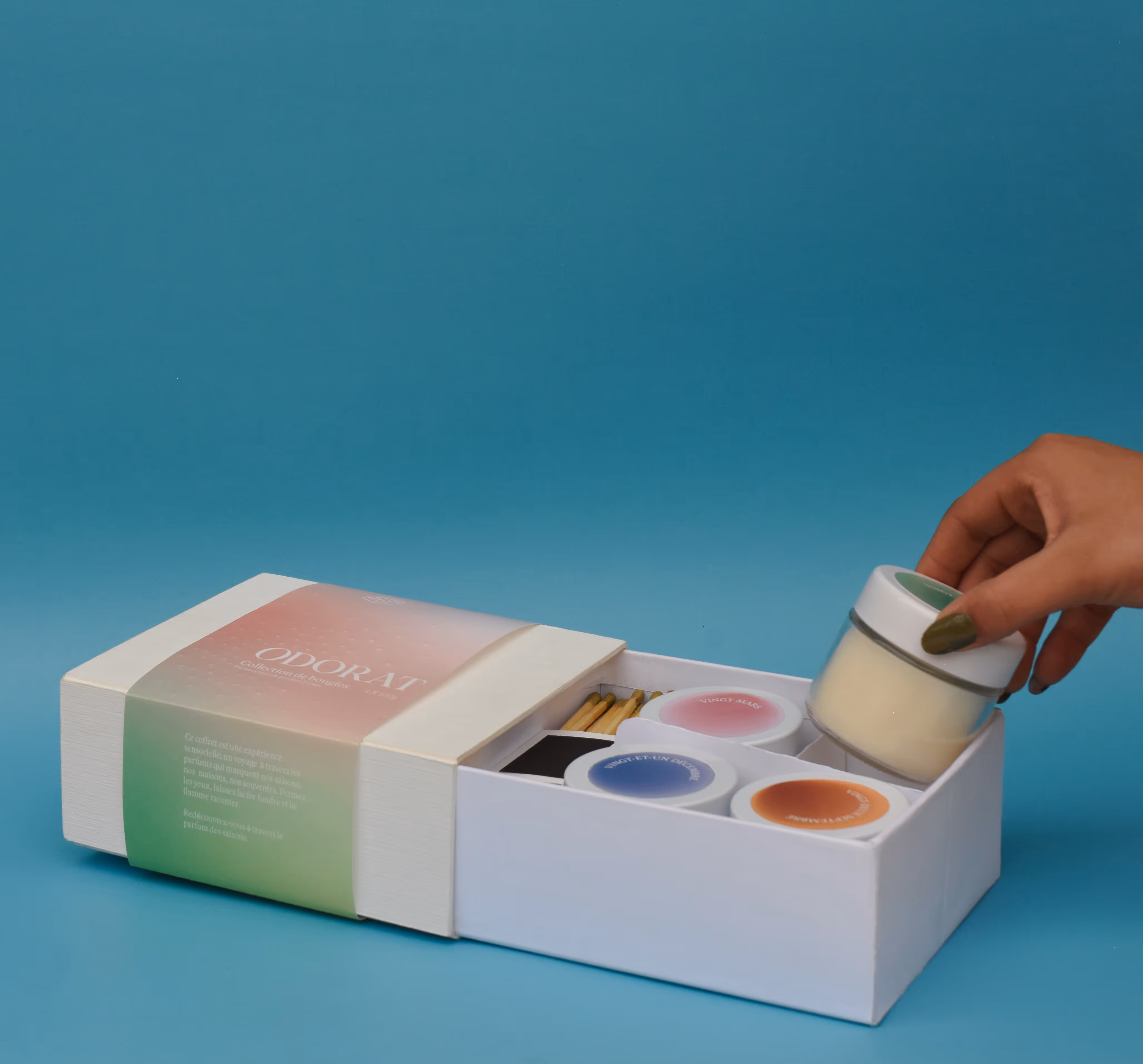 Hand lifting glass candle from an open white box containing four candles and matches on blue background. Credit: May-lee Abel