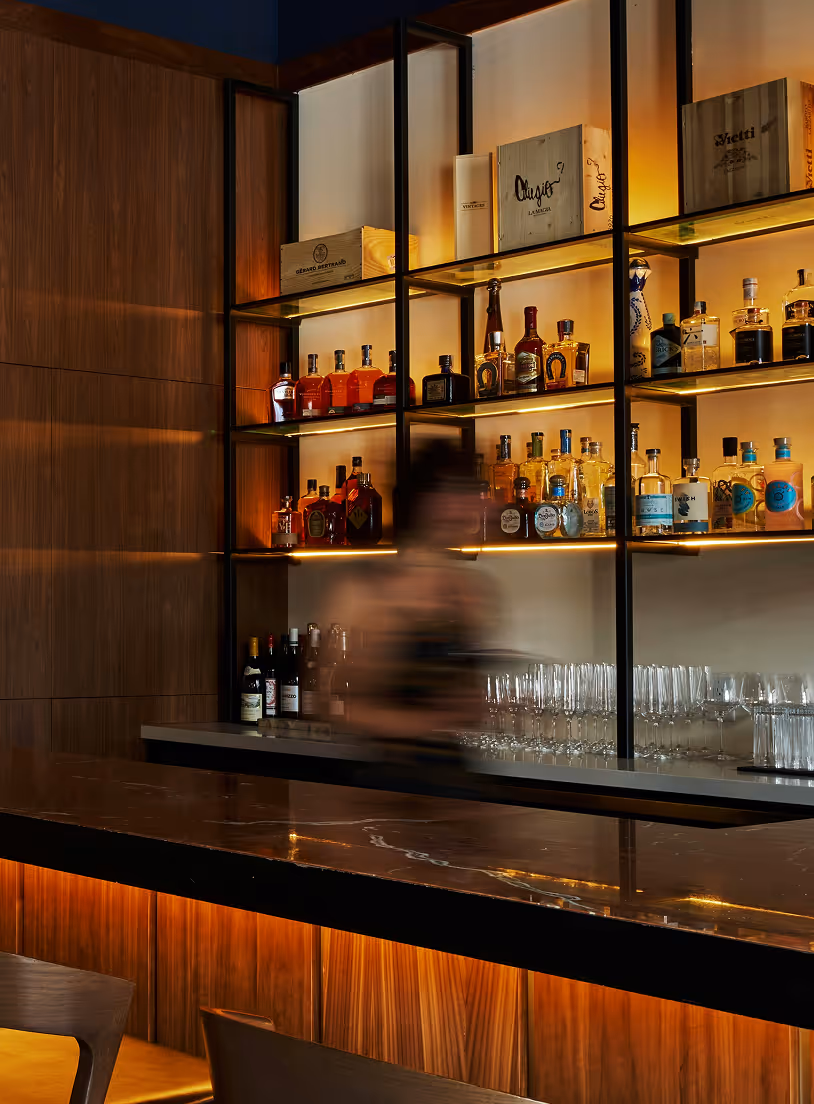 Modern bar with wooden panels, backlit shelves of liquor bottles, glassware, and blurred bartender. Credit: Panos Katsigiannis