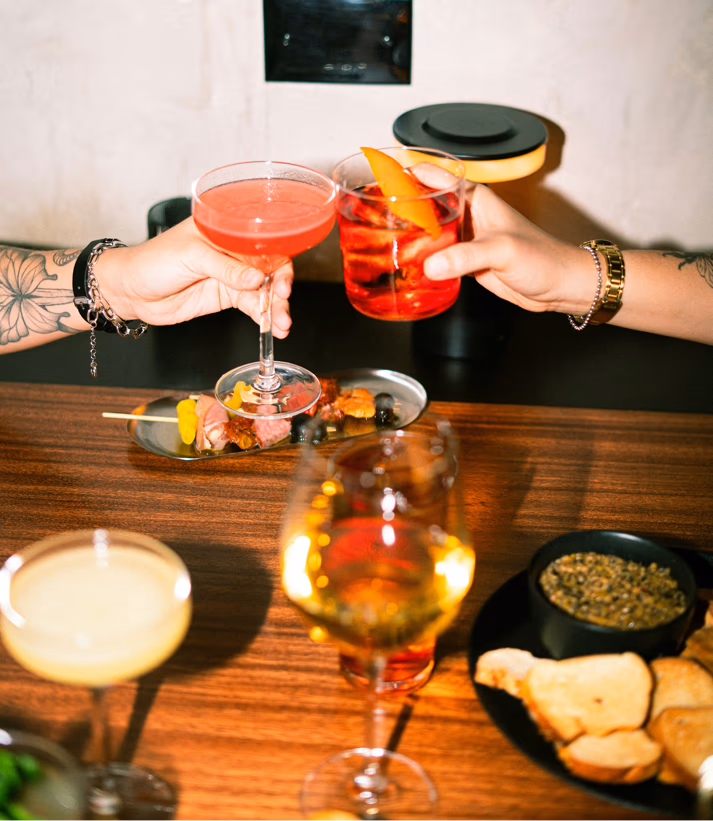 Two tattooed hands clinking cocktails over a table with appetizers and drinks. Credit: Diana Diaz