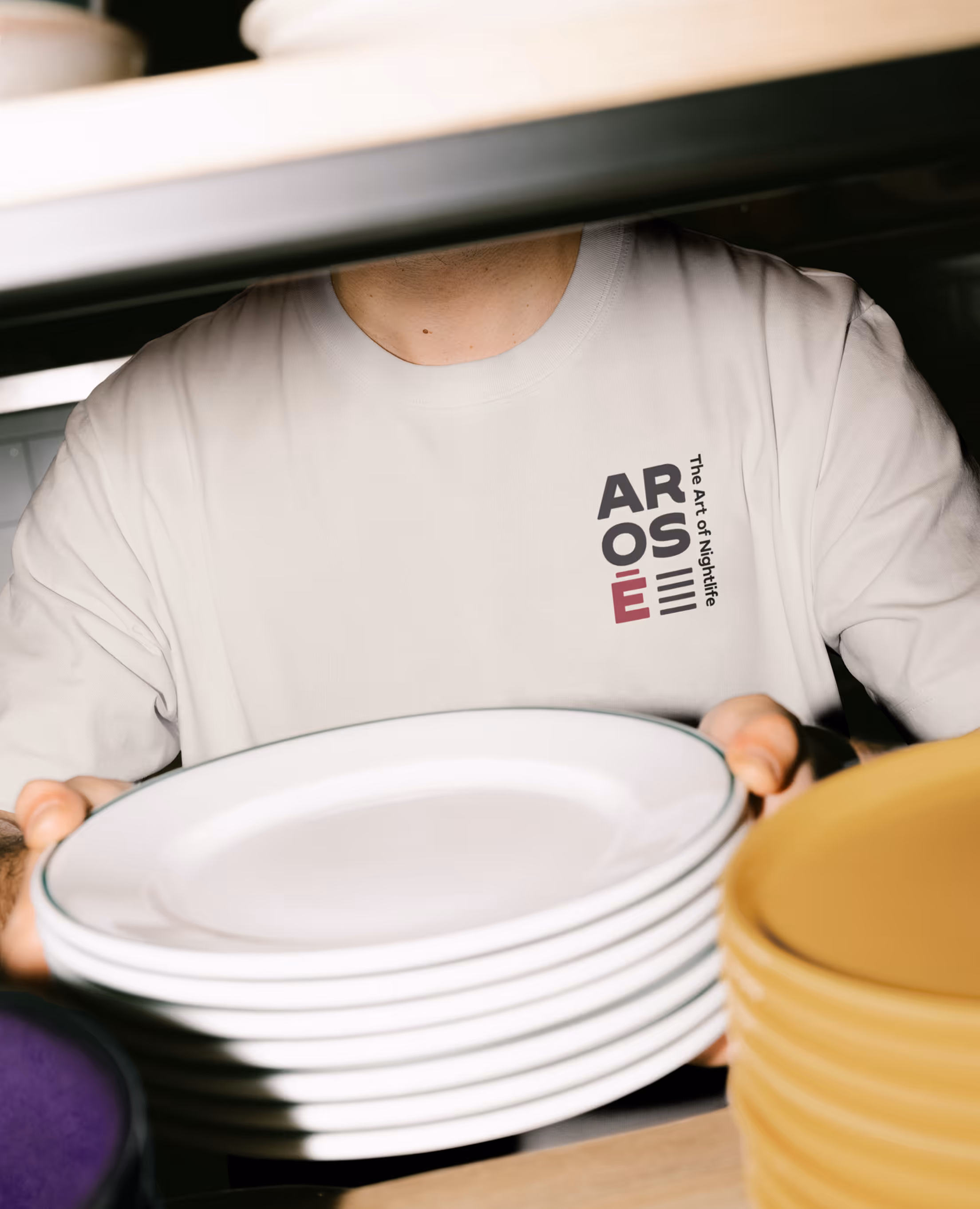 Person holding a stack of white plates wearing a white shirt with ARÔSÉ logo and slogan. Credit: Bendito Mockup