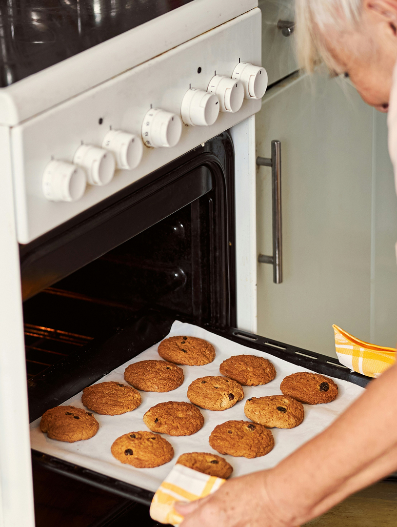 Person removing a tray of freshly baked cookies from a white oven using yellow checkered oven mitts. Credits: Getty Images