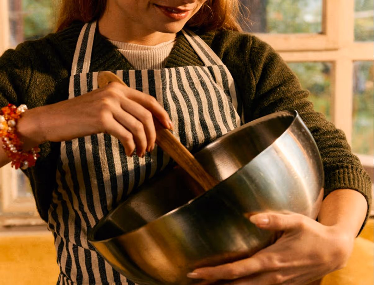 Woman in striped apron stirring contents in a large metal mixing bowl with a wooden spoon. Credit: Daniel Faro
