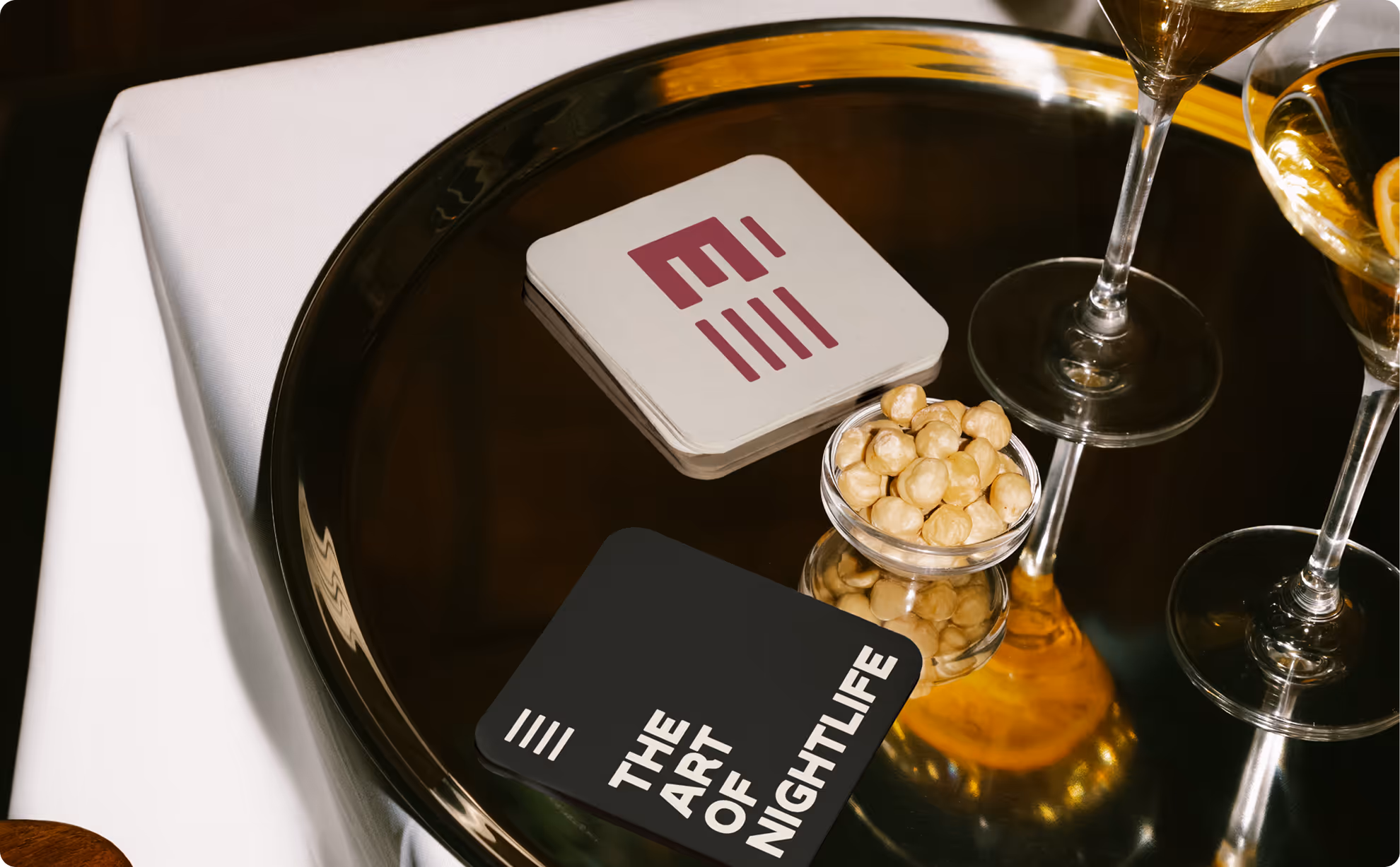 Black tray on white cloth holds coasters reading THE ART OF NIGHTLIFE and a small bowl of nuts. Credit: Bendito Mockup