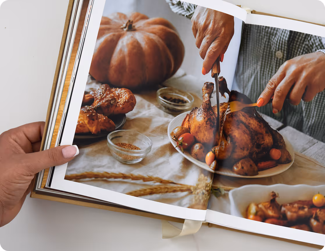 A spread of a book showing a person carving roasted chicken on a plate with vegetables, next to a pumpkin and glass bowls of spices. Credit: May-Lee Abel