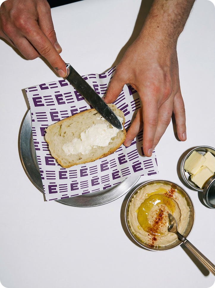 Hands spreading cream cheese on bread next to butter and hummus with olive oil and paprika.