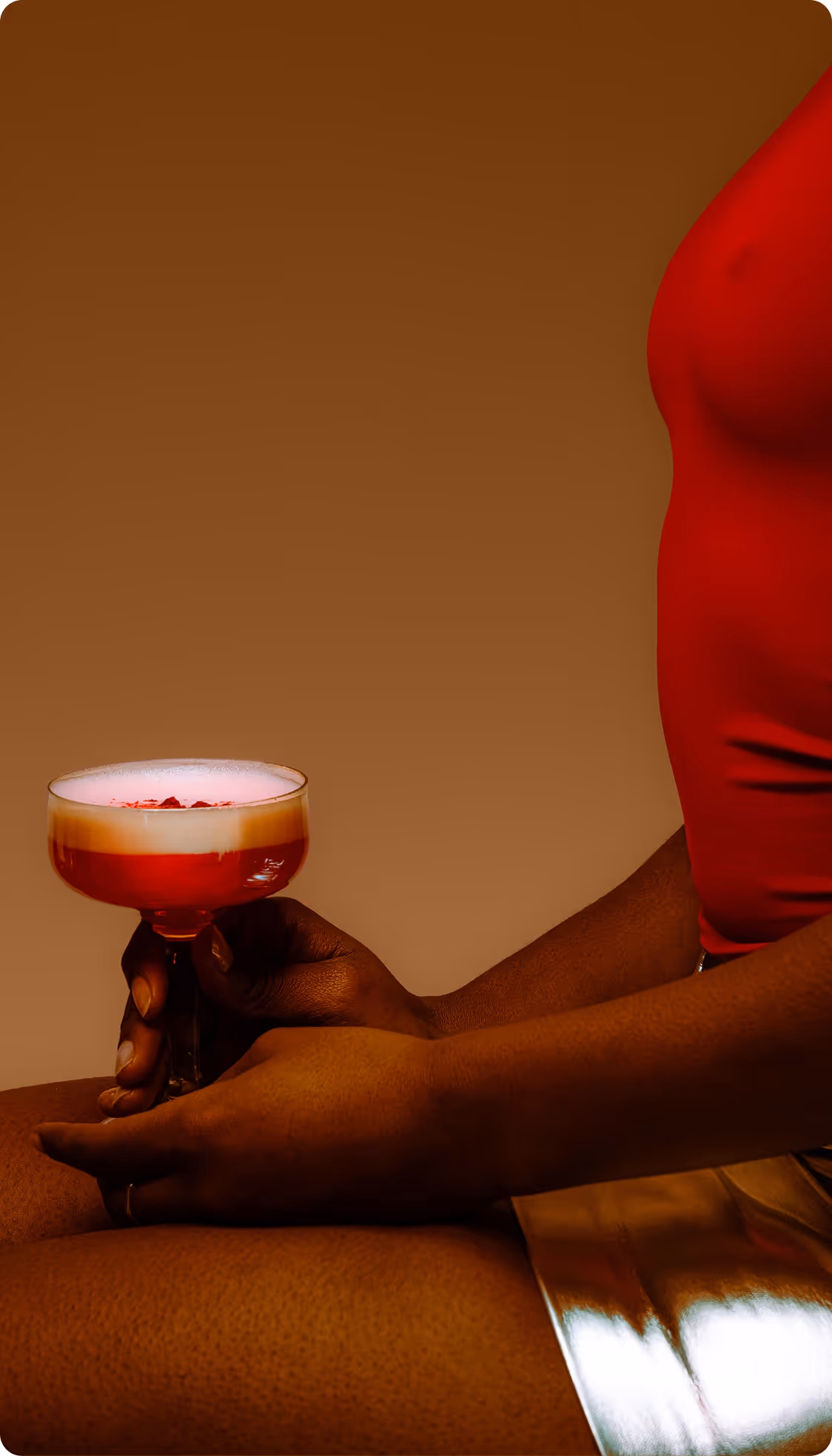 Dark-skinned person in red top and shiny gold shorts holding a cocktail glass with a creamy red drink. Credit: Jelly Luise