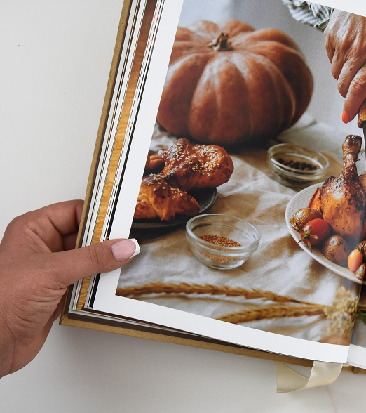Hand holding open a cookbook showing roasted chicken, bread, pumpkin, and small bowls of spices. Credit: May-Lee Abel