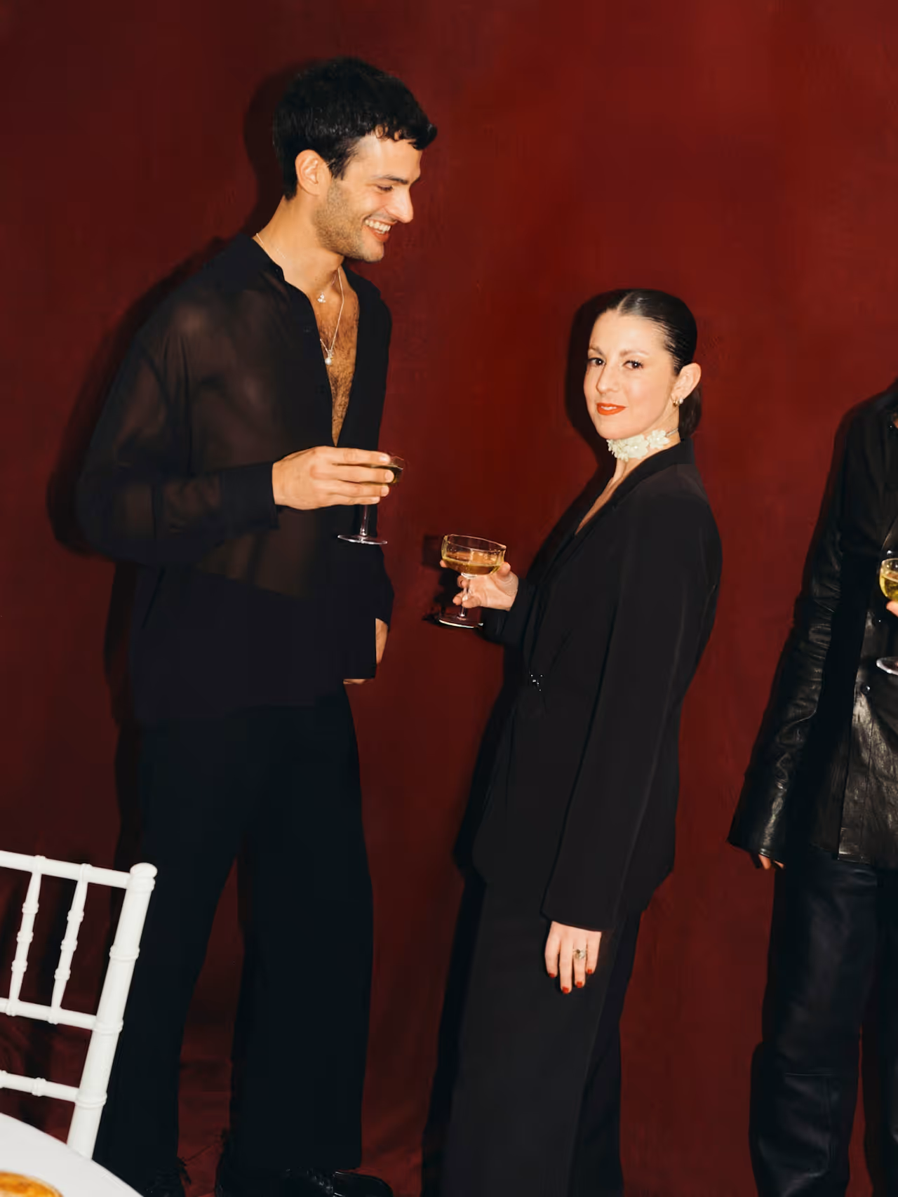 Man and woman dressed in black holding wine glasses, standing against a red background. Credit: Pali Mendez