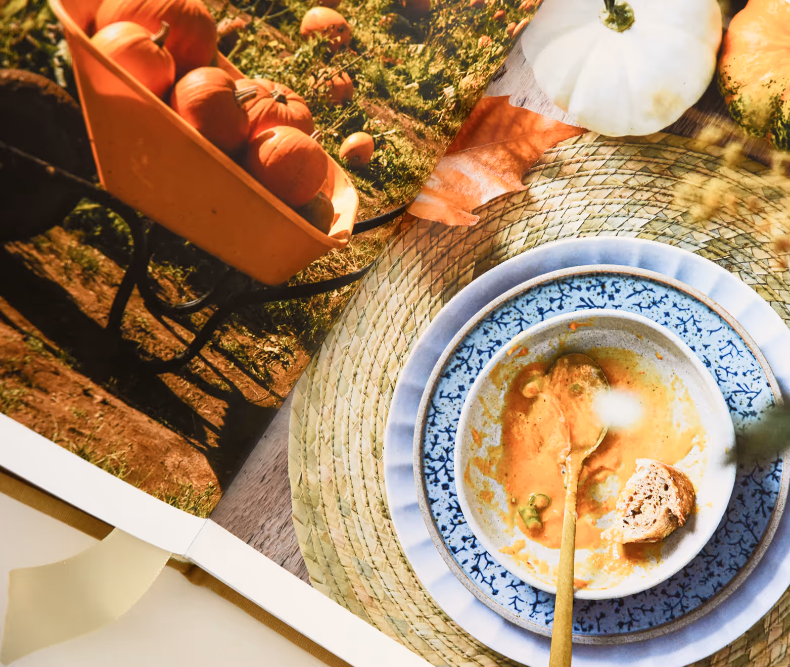 Cookbook spread showing empty bowl with spoon and bread on plates next to a photo of pumpkins in a wheelbarrow. Credit: May-Lee Abel