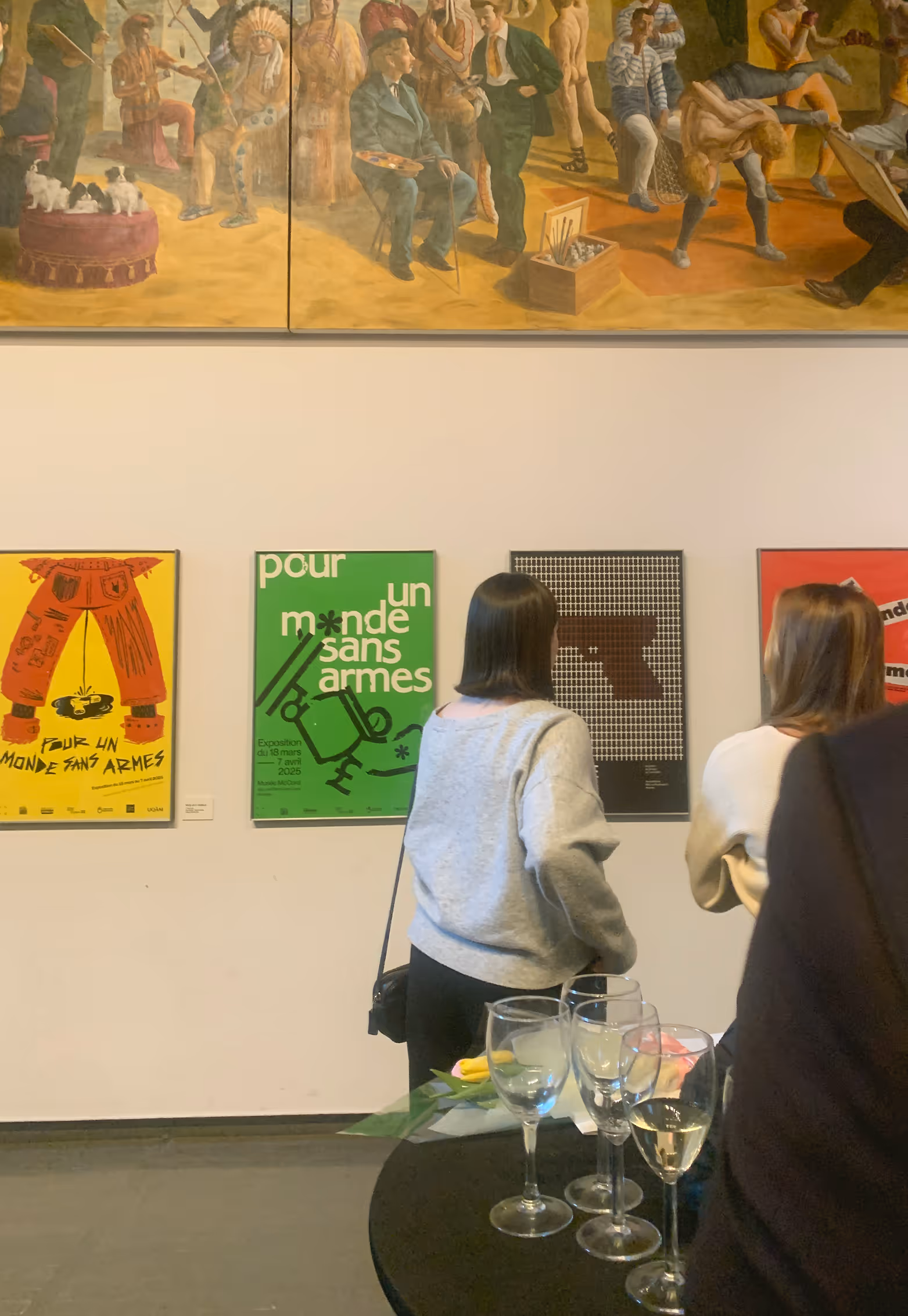 Two women view art posters on a white wall with a mural above and a table with glasses in front. Credit: May-Lee Abel
