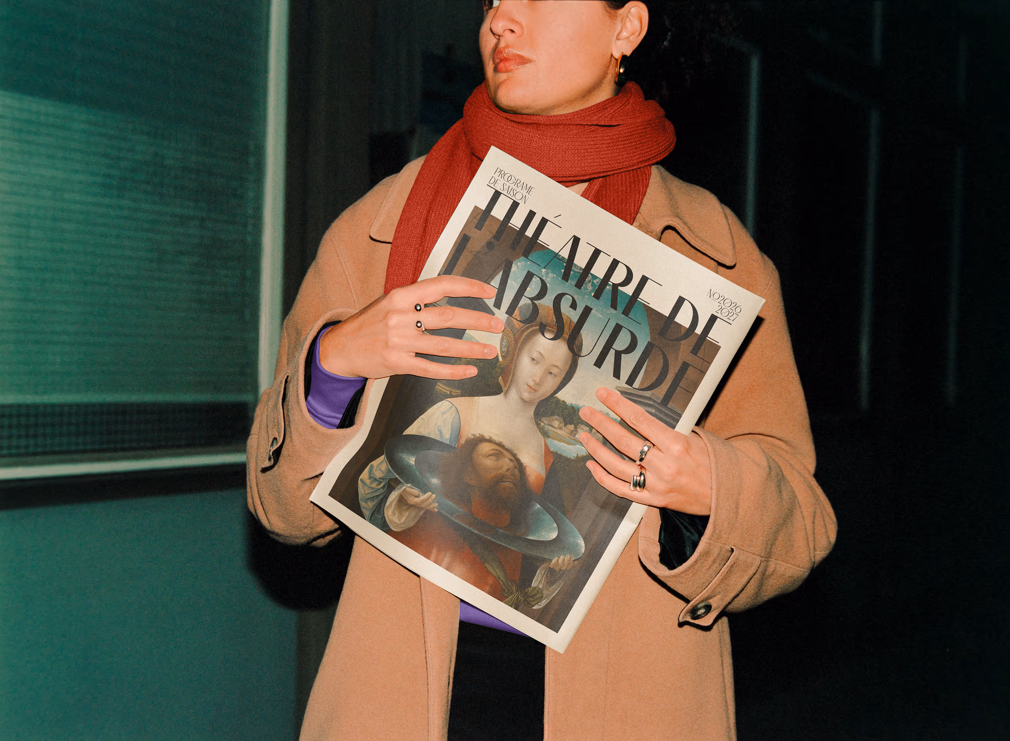 Person wearing a tan coat and red scarf holding a magazine titled Théâtre de l'Absurde at night. Credit: Mockup Maison