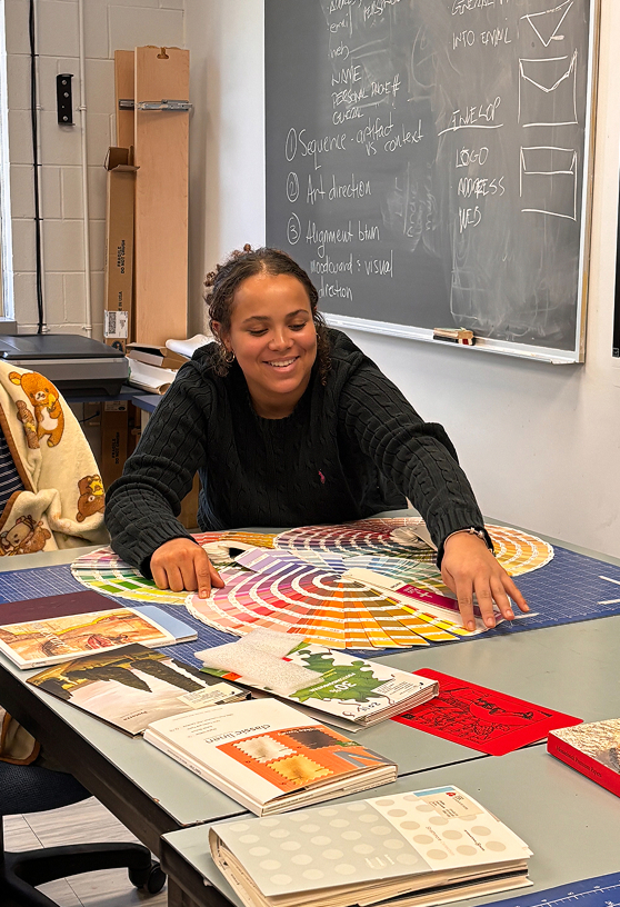 Smiling woman sitting at a table with color swatches and design books in a classroom. Credit: Xhalia Martineau