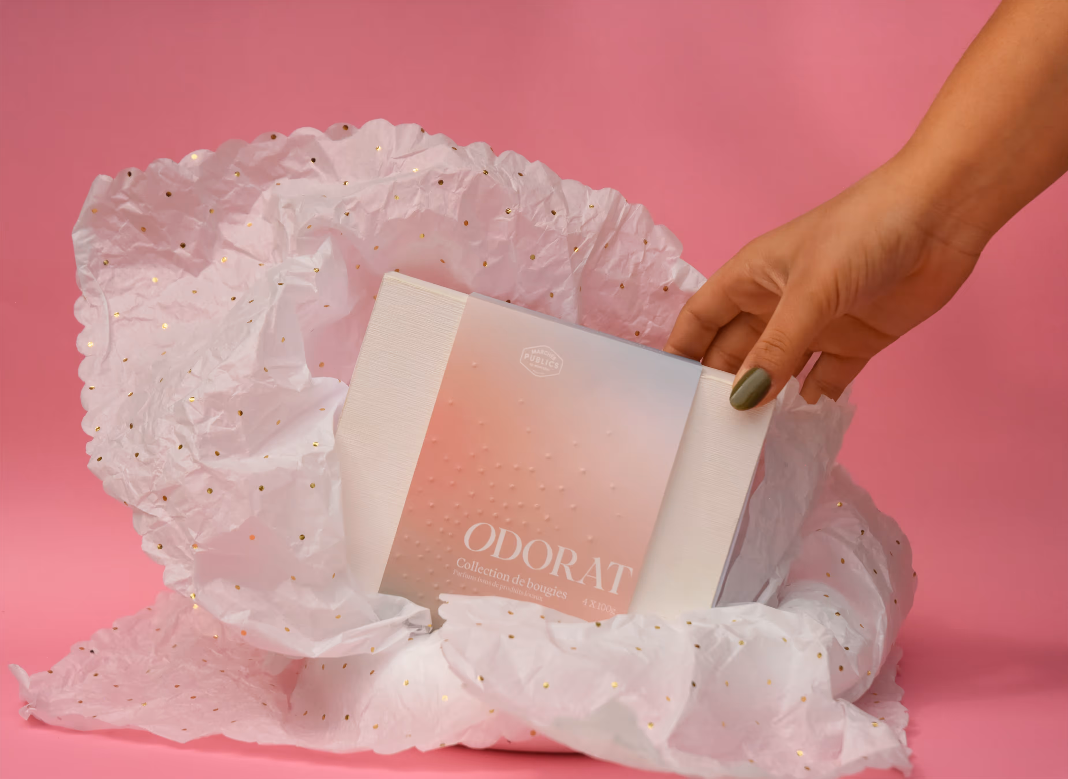 Hand with green nail polish holding a boxed Odorat candle set wrapped in white tissue on pink background. Credit: May-Lee Abel